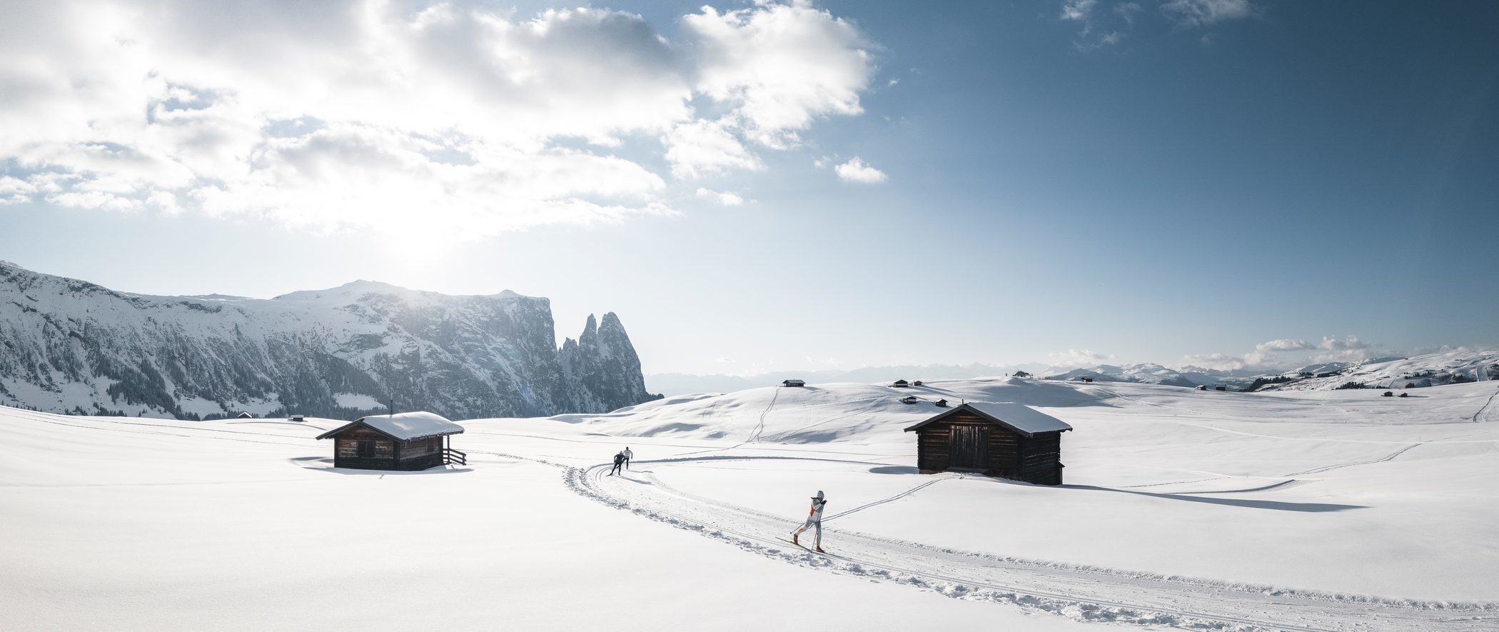 A winter panorama of the Seiser Alm, featuring rustic alpine huts and cross-country ski trails under a blue sky.