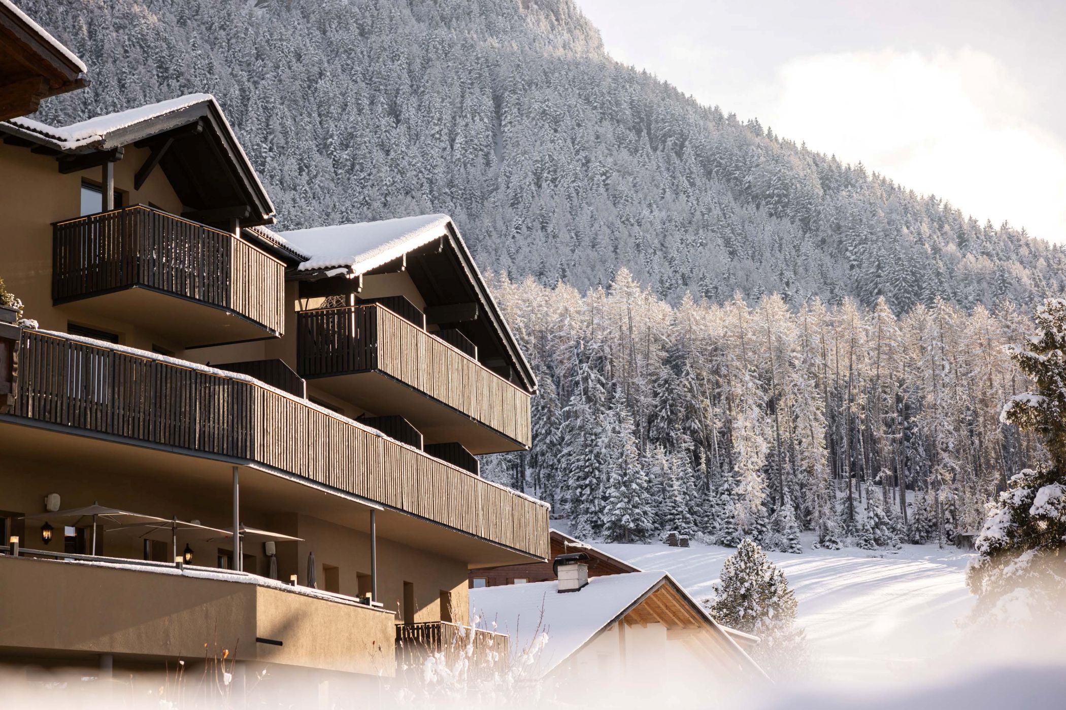 Das Hotel Baumwirt in Kastelruth mit verschneiten Balkonen und winterlichem Wald im Hintergrund.
